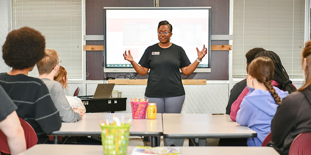 Teacher walking down the aisle between rows of chairs in middle-school classroom.