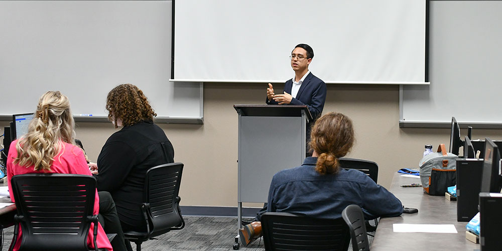 Student giving a presentation at a podium in front of class of students in a computer lab.