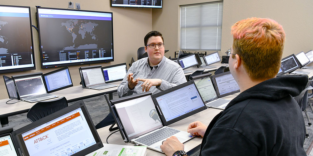 Student in the foreground on a computer receiving help from a student at the next row of computers.