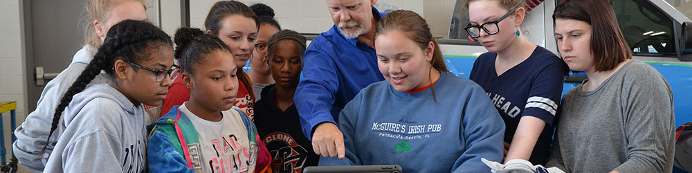 Students inside the automotive lab gathered around a tablet on top of a tool box as the instructor points at the screen.
