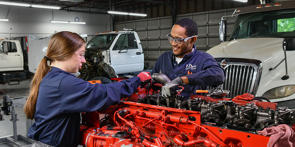 Two students working together to troubleshoot a problem on a diesel engine inside a shop with large trucks in the background.