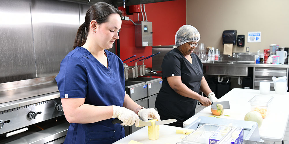 Students in kitchen wearing aprons slicing fruit.