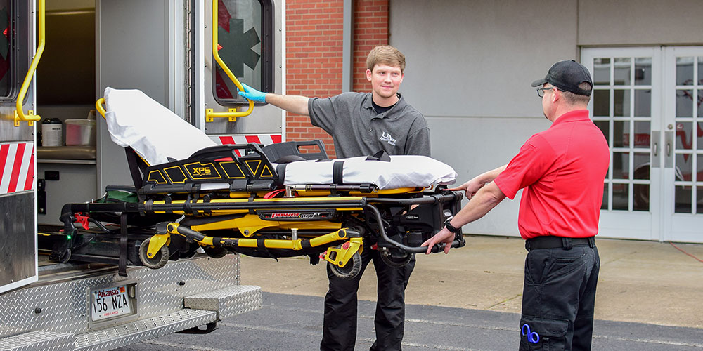 Students at the back of an ambulance learning how to remove and operate a wheeled stetcher.