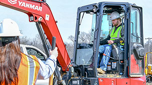 Student operating mini-excavator while student in the foreground watch.