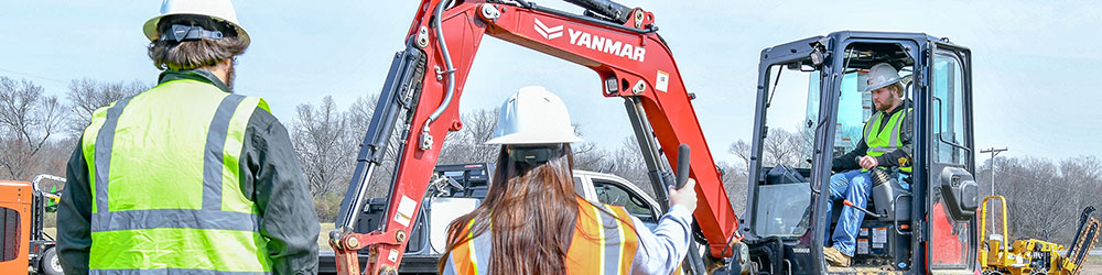 Student operating mini-excavator while two students in the foreground watch.