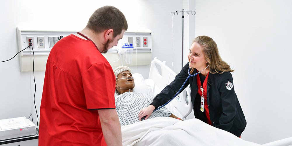 One student checks abdomen of a sim-man with a stethoscope while another student watches.