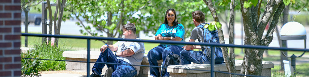 Smiling student holding backpack standing outside buildings with students walking past in background.