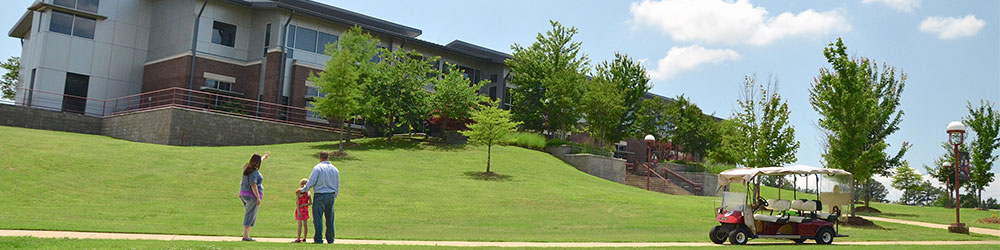 Father and young daughter standing in grass outside University Center as tour guide gestures toward building.